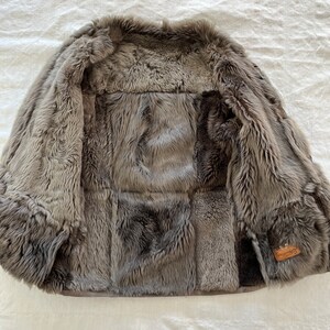 Puede incluir: Una chaqueta de piel de oveja gris con ribete de cuero marr&oacute;n y una peque&ntilde;a etiqueta de cuero marr&oacute;n que dice "Shepherds". La chaqueta est&aacute; abierta y se ve el interior.