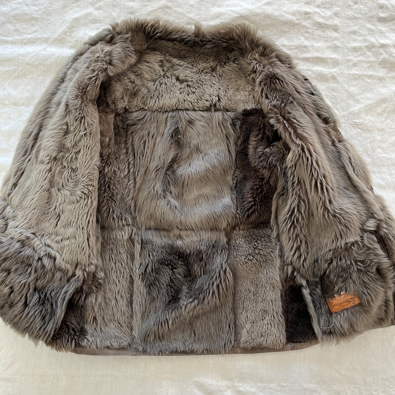 Puede incluir: Una chaqueta de piel de oveja gris con ribete de cuero marr&oacute;n y una peque&ntilde;a etiqueta de cuero marr&oacute;n que dice "Shepherds". La chaqueta est&aacute; abierta y se ve el interior.