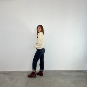 May include: A woman wearing a white sweater with black floral embroidery, dark wash jeans, and burgundy Doc Martens boots.