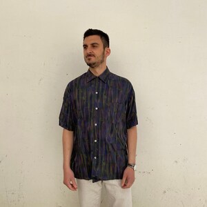 May include: A man wearing a short-sleeved button-down shirt with a dark blue, green, and purple abstract print. The shirt has a single chest pocket and a button-up collar.