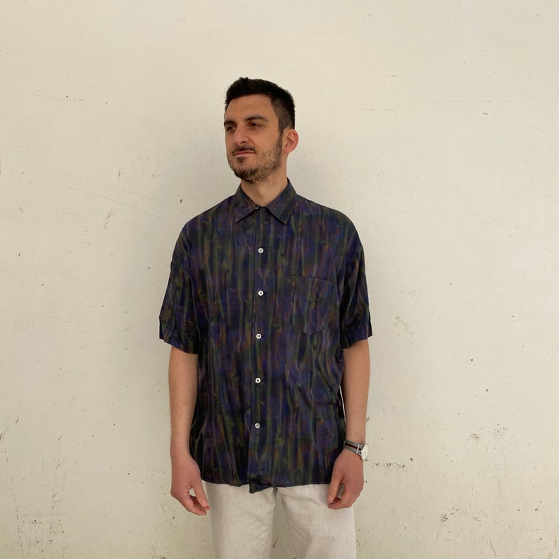May include: A man wearing a short-sleeved button-down shirt with a dark blue, green, and purple abstract print. The shirt has a single chest pocket and a button-up collar.