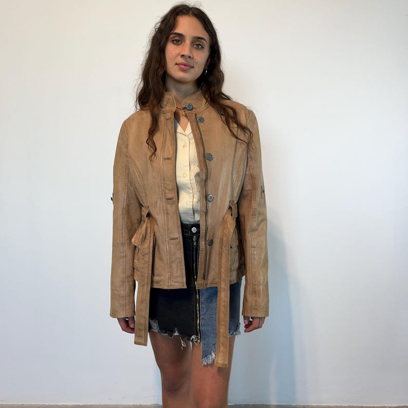 K&ouml;nnte beinhalten: Eine braune Lederjacke mit G&uuml;rtel und Knopfleiste. Die Jacke hat einen Used-Look und wird von einer Person mit langen braunen Haaren getragen.