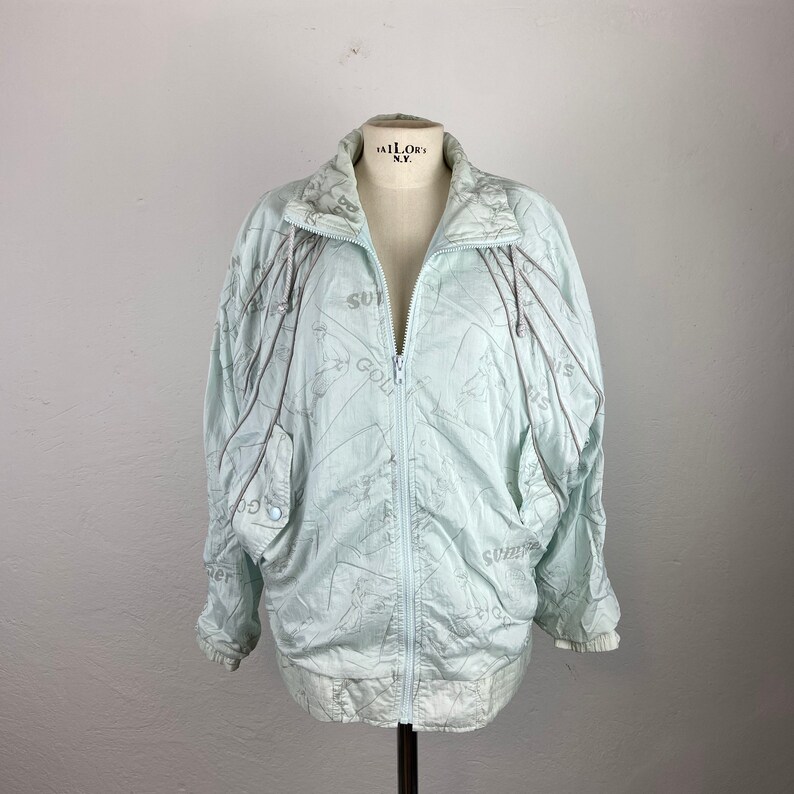 May include: A light blue vintage jacket with a full zipper closure. The jacket has a high collar and features a subtle pattern with the word "Summer" and "Golf" printed on it. The jacket is displayed on a mannequin.