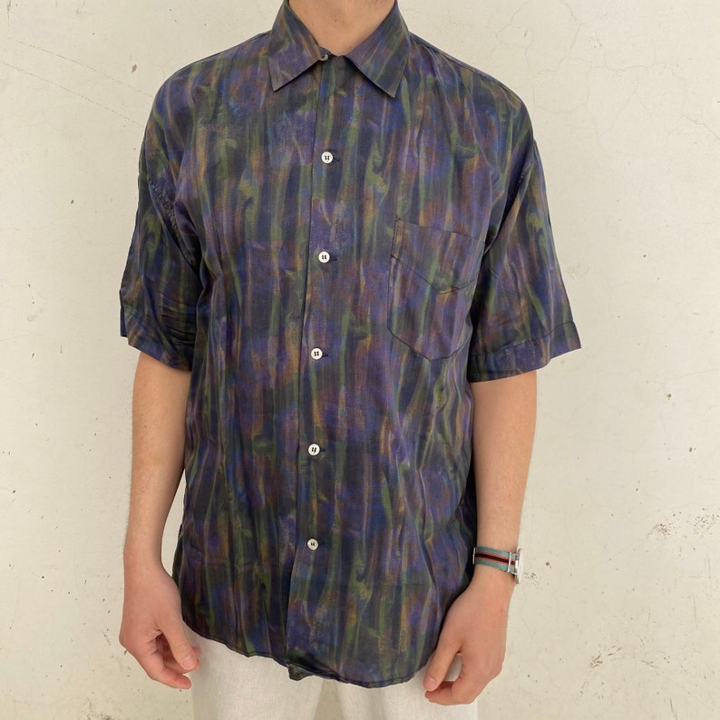 May include: A short-sleeved button-down shirt with a dark blue, green, and purple abstract print. The shirt has a single chest pocket.