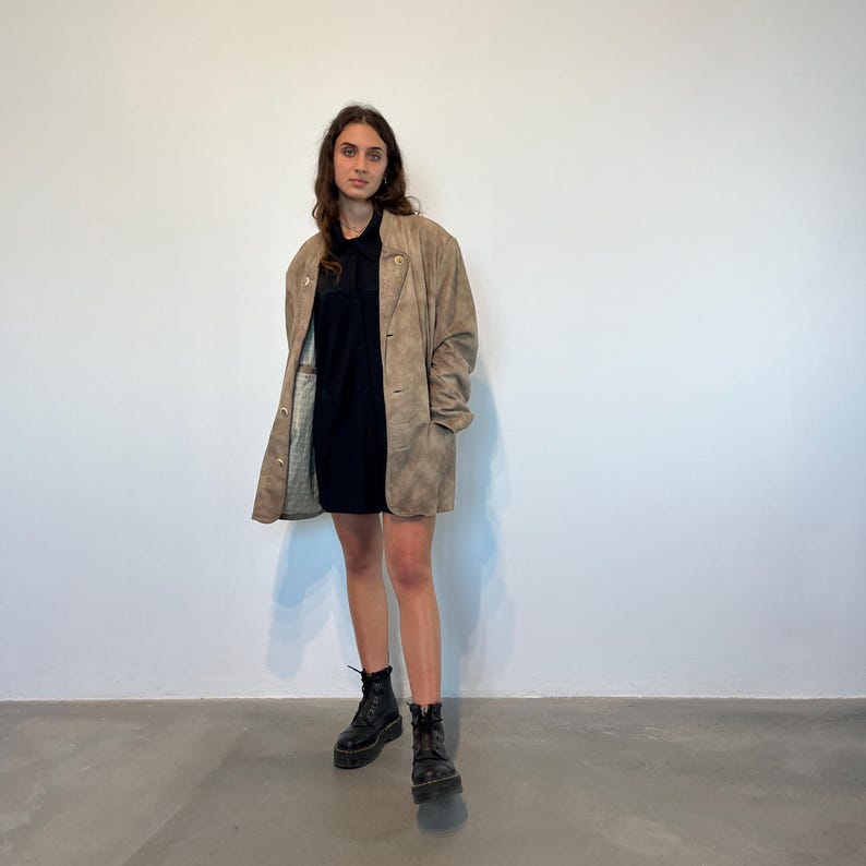 May include: A brown suede jacket with a button closure and a black collared shirt underneath. The person is wearing black boots.