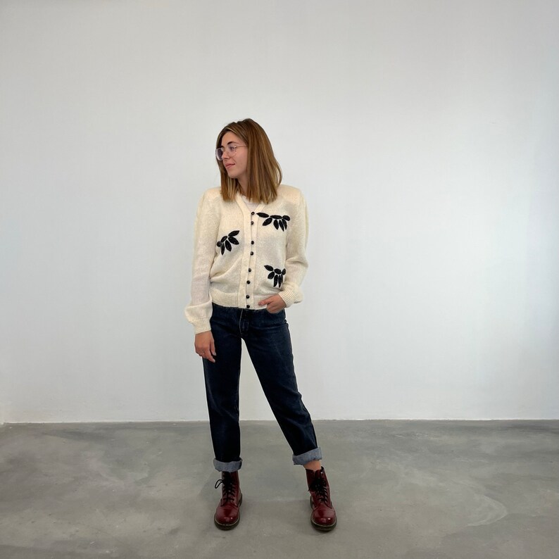 May include: A white cardigan with black floral embroidery. The cardigan is worn over a pair of dark blue jeans. The person is wearing brown boots.