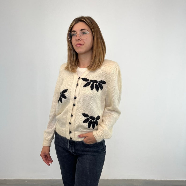 May include: A white cardigan with black floral embroidery and button fastening. The cardigan is made of a soft, fuzzy material.