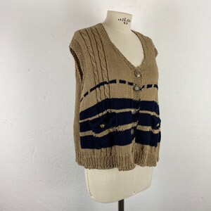 K&ouml;nnte beinhalten: Beige und marineblaue Strickweste mit V-Ausschnitt und Knopfverschluss. Die Weste hat marineblaue horizontale Streifen und kleine Taschen. Die Weste wird auf einer Schneiderpuppe pr&auml;sentiert.
