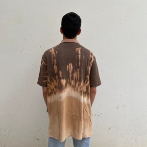 K&ouml;nnte beinhalten: Ein braunes Poloshirt mit Batik-Effekt, das von Dunkelbraun nach Hellbraun &uuml;bergeht. Das Shirt hat kurze &Auml;rmel und einen Kragen. Die Person tr&auml;gt eine hellblaue Jeans.