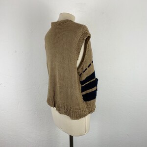 K&ouml;nnte beinhalten: Beige Strickweste mit marineblauen Streifen an der Seite. Die Weste hat einen runden Ausschnitt und einen gewellten Saum. Das &auml;rmellose Design wird auf einer Schaufensterpuppe pr&auml;sentiert. Die Weste ist aus weichem, strukturiertem Garn gefertigt.