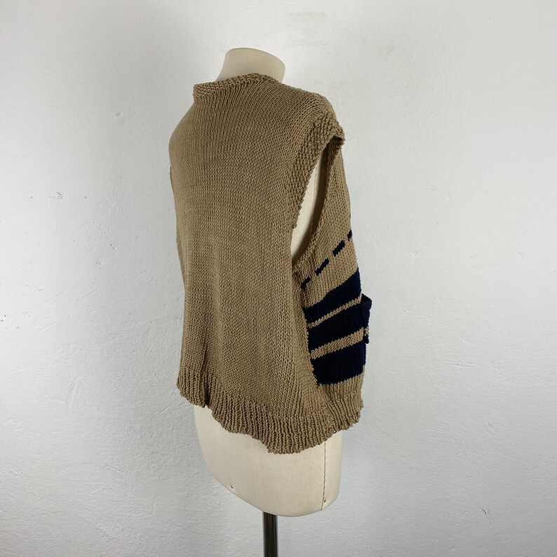 K&ouml;nnte beinhalten: Beige Strickweste mit marineblauen Streifen an der Seite. Die Weste hat einen runden Ausschnitt und einen gewellten Saum. Das &auml;rmellose Design wird auf einer Schaufensterpuppe pr&auml;sentiert. Die Weste ist aus weichem, strukturiertem Garn gefertigt.