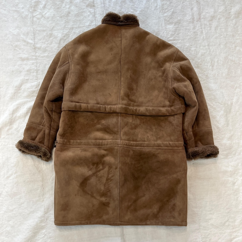 May include: A brown sheepskin coat with a fur collar and a button closure. The coat has a long length and a relaxed fit.
