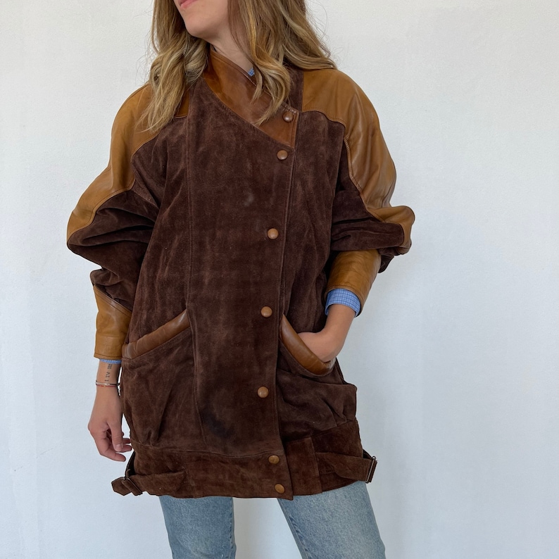 May include: A brown suede jacket with brown leather sleeves. The jacket has a button closure and two pockets.