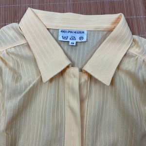 May include: A pale yellow, striped, long-sleeved shirt with a button-up front and a collar. The shirt has a label that reads "100% POLYESTER" and includes care instructions and a size 42.