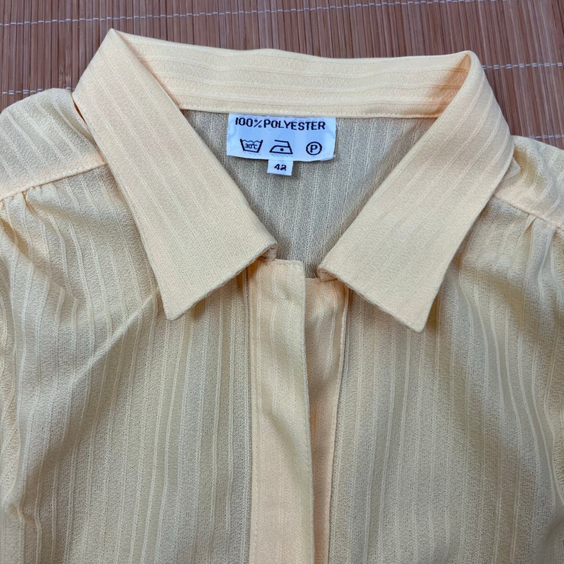 May include: A pale yellow, striped, long-sleeved shirt with a button-up front and a collar. The shirt has a label that reads "100% POLYESTER" and includes care instructions and a size 42.