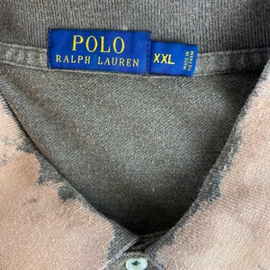 K&ouml;nnte beinhalten: Ein graues und beiges Polo Ralph Lauren Hemd. Das Hemd hat einen Kragen und einen Knopfverschluss. Das Etikett lautet "POLO RALPH LAUREN" in goldener Schrift auf blauem Hintergrund. Das Hemd ist in der Gr&ouml;&szlig;e XXL.