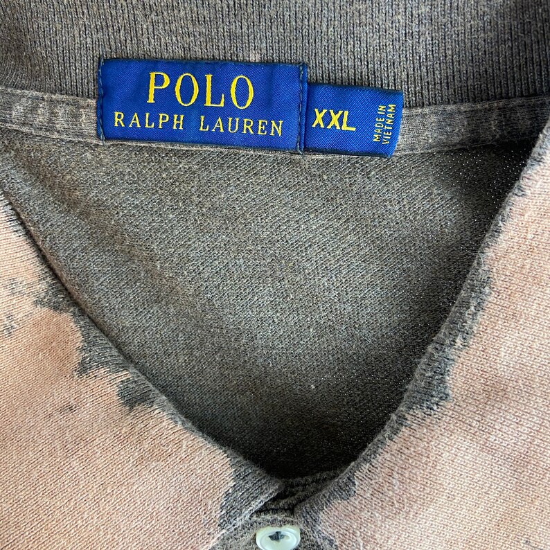 K&ouml;nnte beinhalten: Ein graues und beiges Polo Ralph Lauren Hemd. Das Hemd hat einen Kragen und einen Knopfverschluss. Das Etikett lautet "POLO RALPH LAUREN" in goldener Schrift auf blauem Hintergrund. Das Hemd ist in der Gr&ouml;&szlig;e XXL.