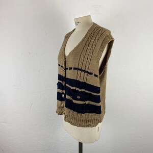 K&ouml;nnte beinhalten: Beige Strickweste mit marineblauen Streifen und silbernen Kn&ouml;pfen. Die &auml;rmellose Weste hat einen V-Ausschnitt und zwei Taschen. Das Strickmuster umfasst Zopfmusterdetails an den Schultern. Die Weste wird auf einer Schaufensterpuppe pr&auml;sentiert.