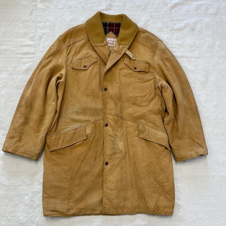 May include: A long, tan leather jacket with a plaid lining on the collar. The jacket has a button closure, two large flap pockets on the front, and two smaller pockets on the sides.