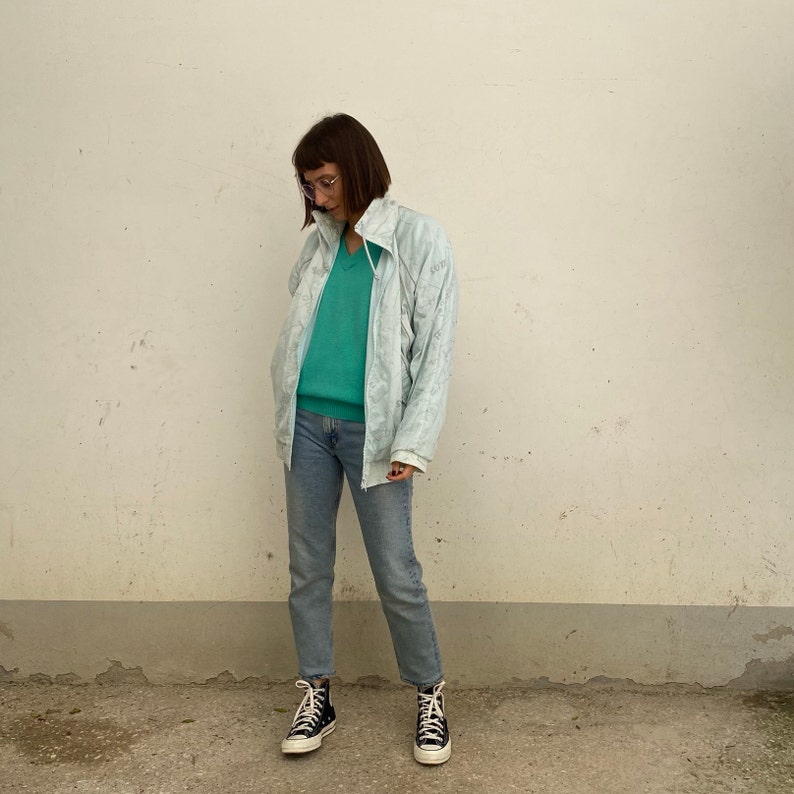 May include: A light blue zip-up jacket with a subtle pattern, paired with a teal v-neck sweater and light wash jeans. The outfit is completed with black high-top sneakers. The jacket has a high collar and a zipper closure.