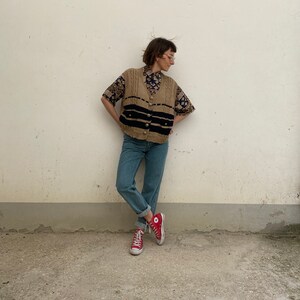 K&ouml;nnte beinhalten: Eine Person tr&auml;gt eine beige Strickweste mit schwarzen Streifen und goldenen Kn&ouml;pfen &uuml;ber einem gemusterten Kurzarmhemd. Die Person tr&auml;gt hellblaue Jeans und rote High-Top-Sneakers.