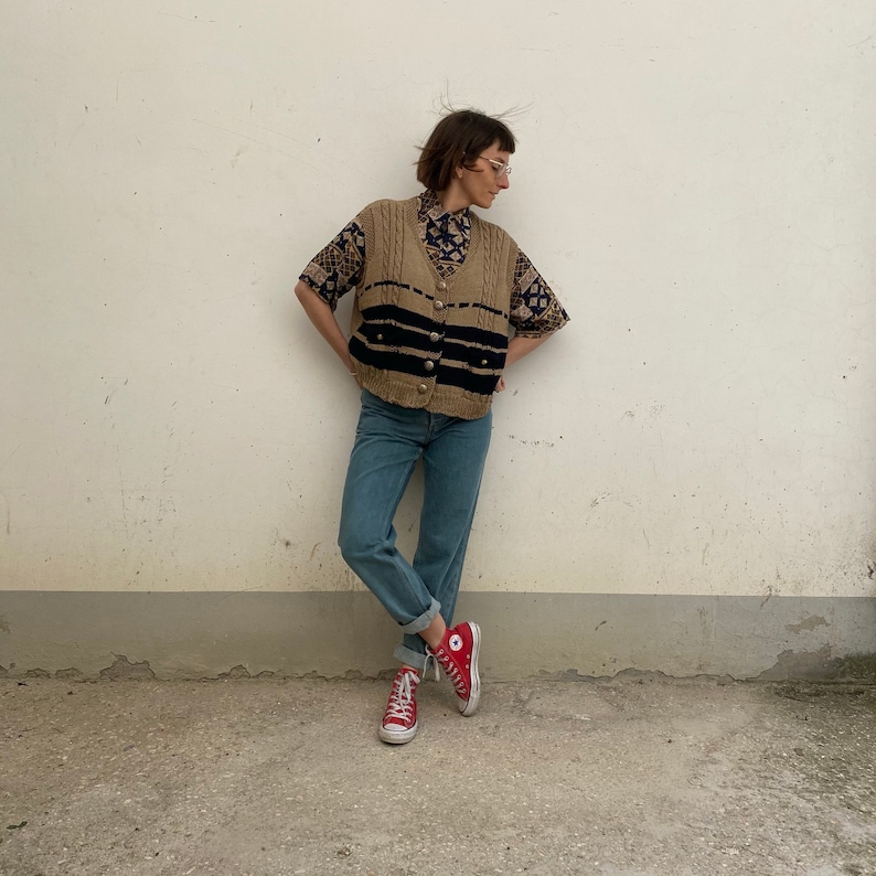 K&ouml;nnte beinhalten: Eine Person tr&auml;gt eine beige Strickweste mit schwarzen Streifen und goldenen Kn&ouml;pfen &uuml;ber einem gemusterten Kurzarmhemd. Die Person tr&auml;gt hellblaue Jeans und rote High-Top-Sneakers.