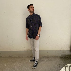 May include: A man wearing a short-sleeved, patterned button-down shirt in shades of blue, green, and black. He is wearing light-wash jeans and black sneakers.