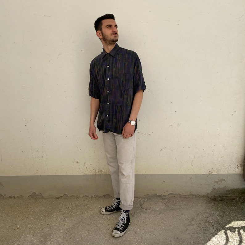 May include: A man wearing a short-sleeved, patterned button-down shirt in shades of blue, green, and black. He is wearing light-wash jeans and black sneakers.