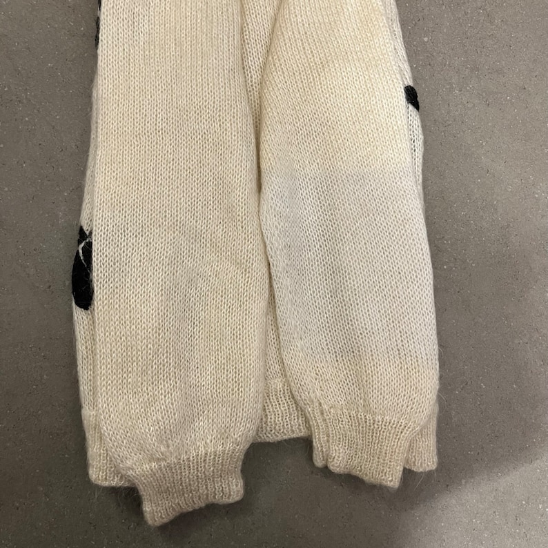 May include: A cream-coloured knitted sweater with black embroidered details. The sweater has a ribbed texture and a loose fit.