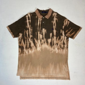 K&ouml;nnte beinhalten: Ein braunes Poloshirt mit einem Bleichmittel-Design. Das Shirt hat einen Kragen, kurze &Auml;rmel und ein kleines blaues Logo auf der Brust. Der Bleicheffekt erzeugt ein hellbraunes und wei&szlig;es Muster.