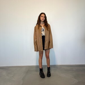 May include: A brown leather jacket with a button-up front and long sleeves. The jacket is worn by a person with long brown hair and is paired with a black mini skirt and black boots.