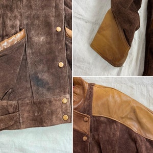 May include: A brown suede jacket with brown leather accents. The jacket has a zipper closure and a stand-up collar. The jacket has a patch pocket on the left chest and two flap pockets at the waist.