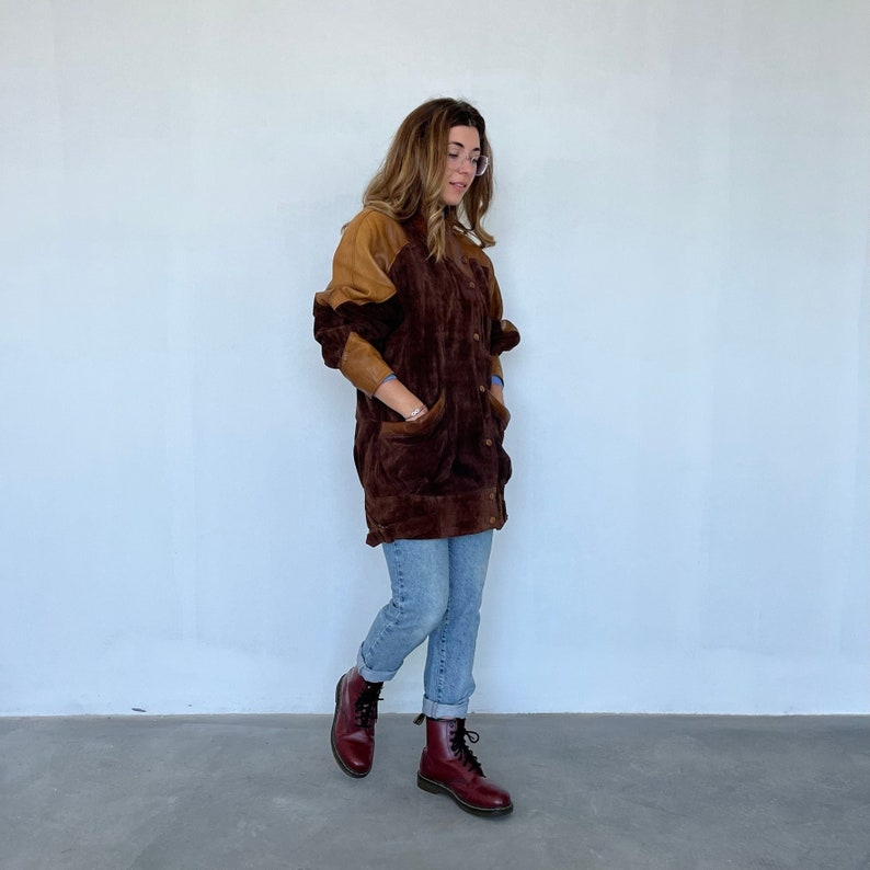 May include: A brown and tan suede jacket with a button closure. The jacket has a long, relaxed fit and is worn with blue jeans and burgundy boots.