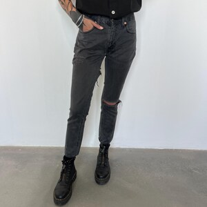 May include: Faded black denim jeans with a distressed knee and thigh detail. The jeans are paired with black platform boots with a front zipper. The model is wearing a black top.