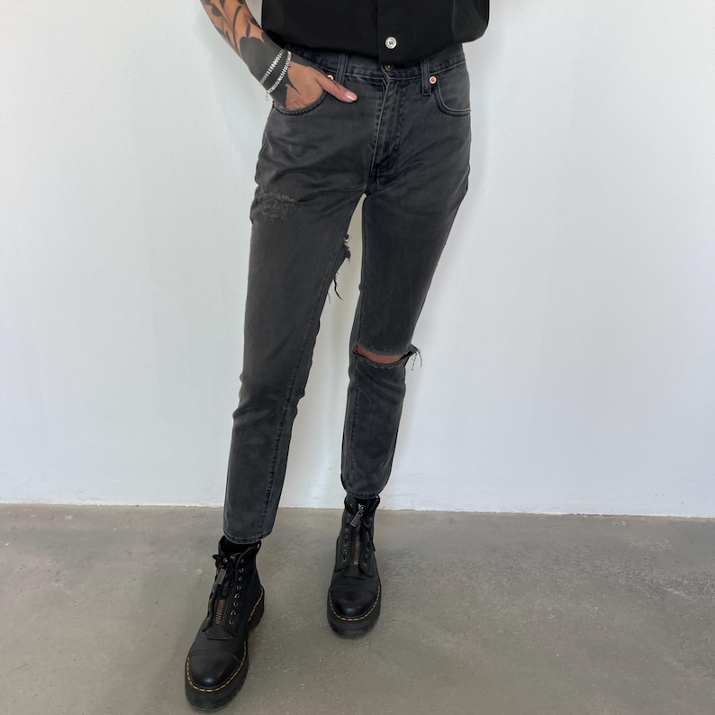 May include: Faded black denim jeans with a distressed knee and thigh detail. The jeans are paired with black platform boots with a front zipper. The model is wearing a black top.