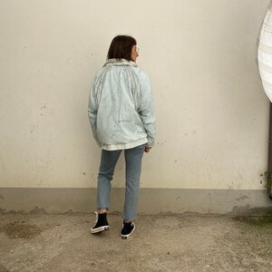 May include: A light blue jacket with a map-like design and the text "SUBSTANCE" and "FEELINGS". The jacket is paired with light blue jeans and black sneakers. The person is standing in front of a white wall.