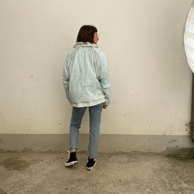 May include: A light blue jacket with a map-like design and the text "SUBSTANCE" and "FEELINGS". The jacket is paired with light blue jeans and black sneakers. The person is standing in front of a white wall.