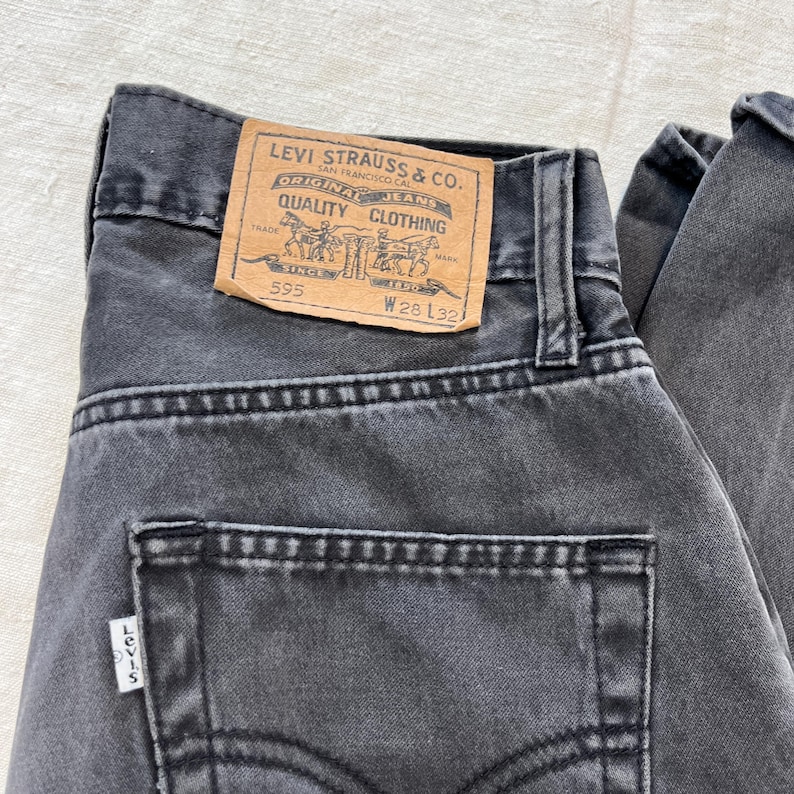 May include: Close-up of a pair of faded black Levi's jeans. The back pocket and the Levi's label are visible. The label reads "LEVI STRAUSS & CO. ORIGINAL JEANS QUALITY CLOTHING" with the numbers "595", "W28 L32", and the Levi's logo.