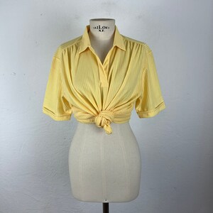 May include: A yellow short-sleeved collared shirt with a ribbed texture. The shirt is tied at the waist and has the text "TAILOR'S N.Y." on the collar.