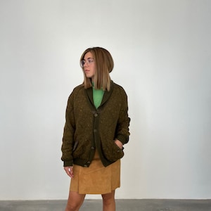 May include: A brown cardigan with a collared neckline and a button closure. The cardigan has a speckled pattern and is worn over a green turtleneck shirt. The person is wearing a brown skirt.