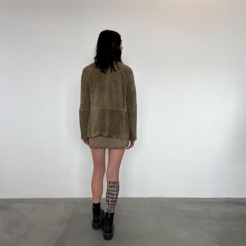 May include: A brown suede jacket with a zip closure and a brown suede mini skirt. The person is wearing black boots.