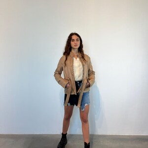 K&ouml;nnte beinhalten: Eine braune Lederjacke mit Knopfleiste und G&uuml;rtel. Die Jacke hat einen Stehkragen und lange &Auml;rmel. Das Model tr&auml;gt darunter ein wei&szlig;es Hemd mit Kn&ouml;pfen und eine Jeans-Shorts.