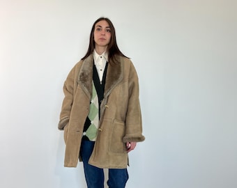 May include: A tan shearling coat with a brown fur collar and cuffs. The coat has two front pockets and gold buttons. The person is wearing a white shirt, a green argyle sweater, and blue jeans.