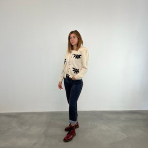May include: A white cardigan with black floral embroidery. The cardigan has a button-up front and long sleeves. The model is wearing the cardigan with blue jeans and brown boots.