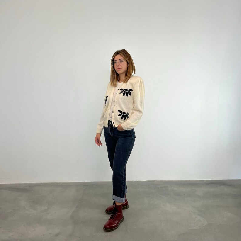 May include: A white cardigan with black floral embroidery. The cardigan has a button-up front and long sleeves. The model is wearing the cardigan with blue jeans and brown boots.