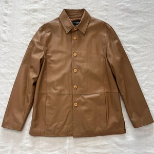 May include: A brown leather jacket with a button-up front and a collar. The jacket has a single pocket on the left side and a label that reads "Gianni Versace".