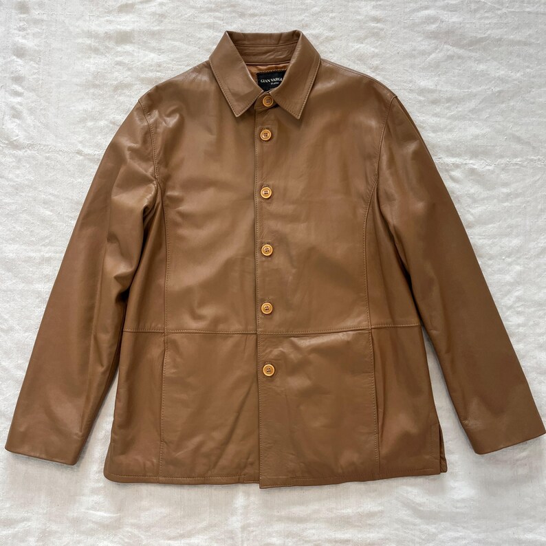 May include: A brown leather jacket with a button-up front and a collar. The jacket has a single pocket on the left side and a label that reads "Gianni Versace".