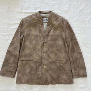 May include: A brown suede jacket with a mandarin collar and four gold buttons. The jacket has two pockets on the front and a label on the inside collar that reads "Trachtenmode".