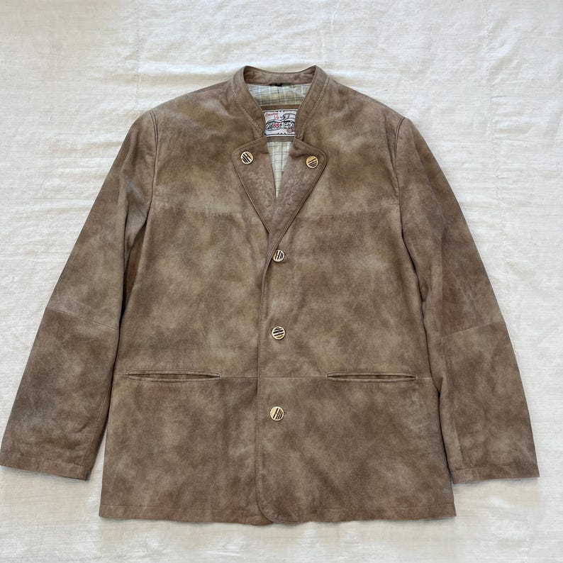 May include: A brown suede jacket with a mandarin collar and four gold buttons. The jacket has two pockets on the front and a label on the inside collar that reads "Trachtenmode".