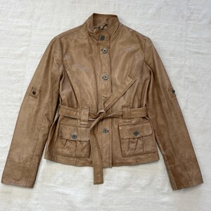 K&ouml;nnte beinhalten: Eine braune Lederjacke mit G&uuml;rtel und Taschen. Die Jacke hat einen Stehkragen und einen Knopfverschluss.